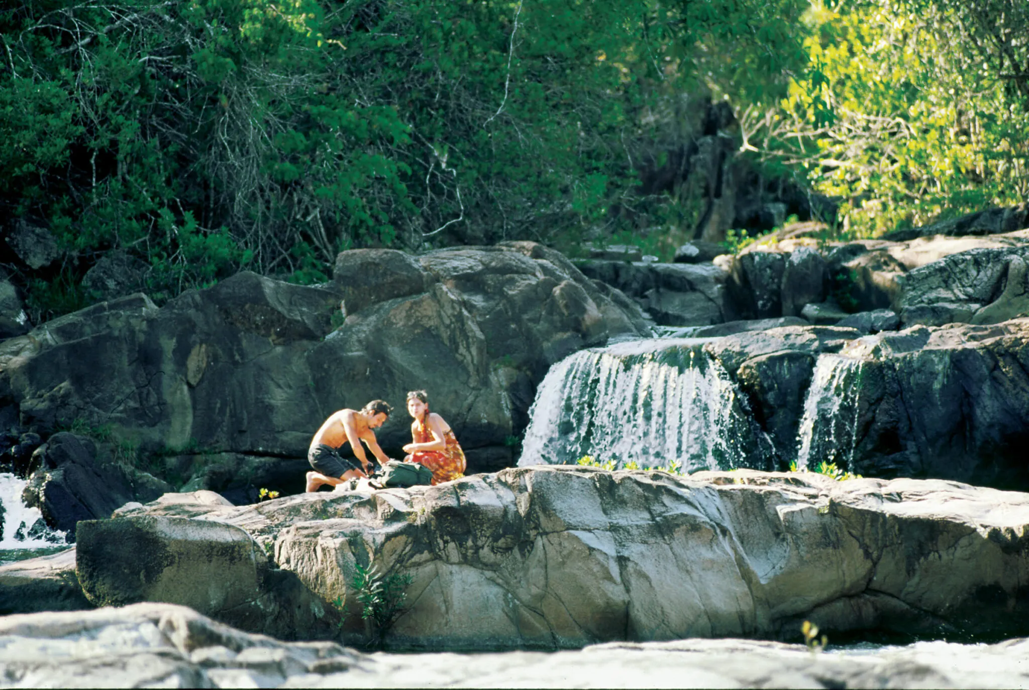 A couple relaxes on a rock formation near a waterfall