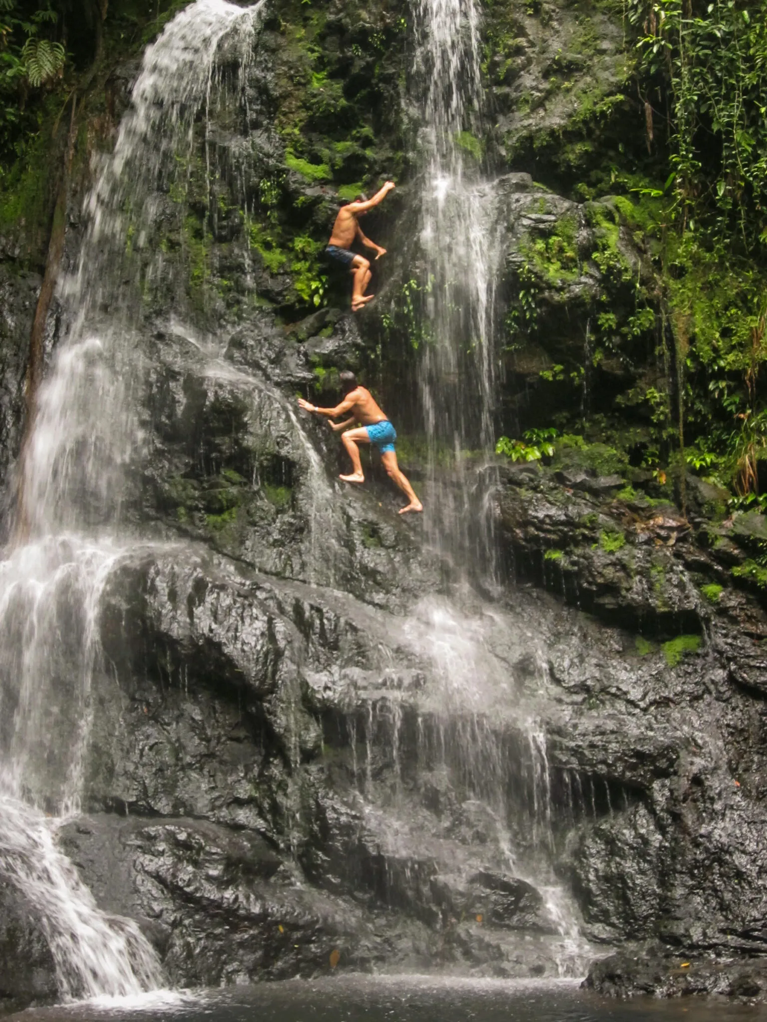 Men in swim trunks climb rocks behind a waterfall