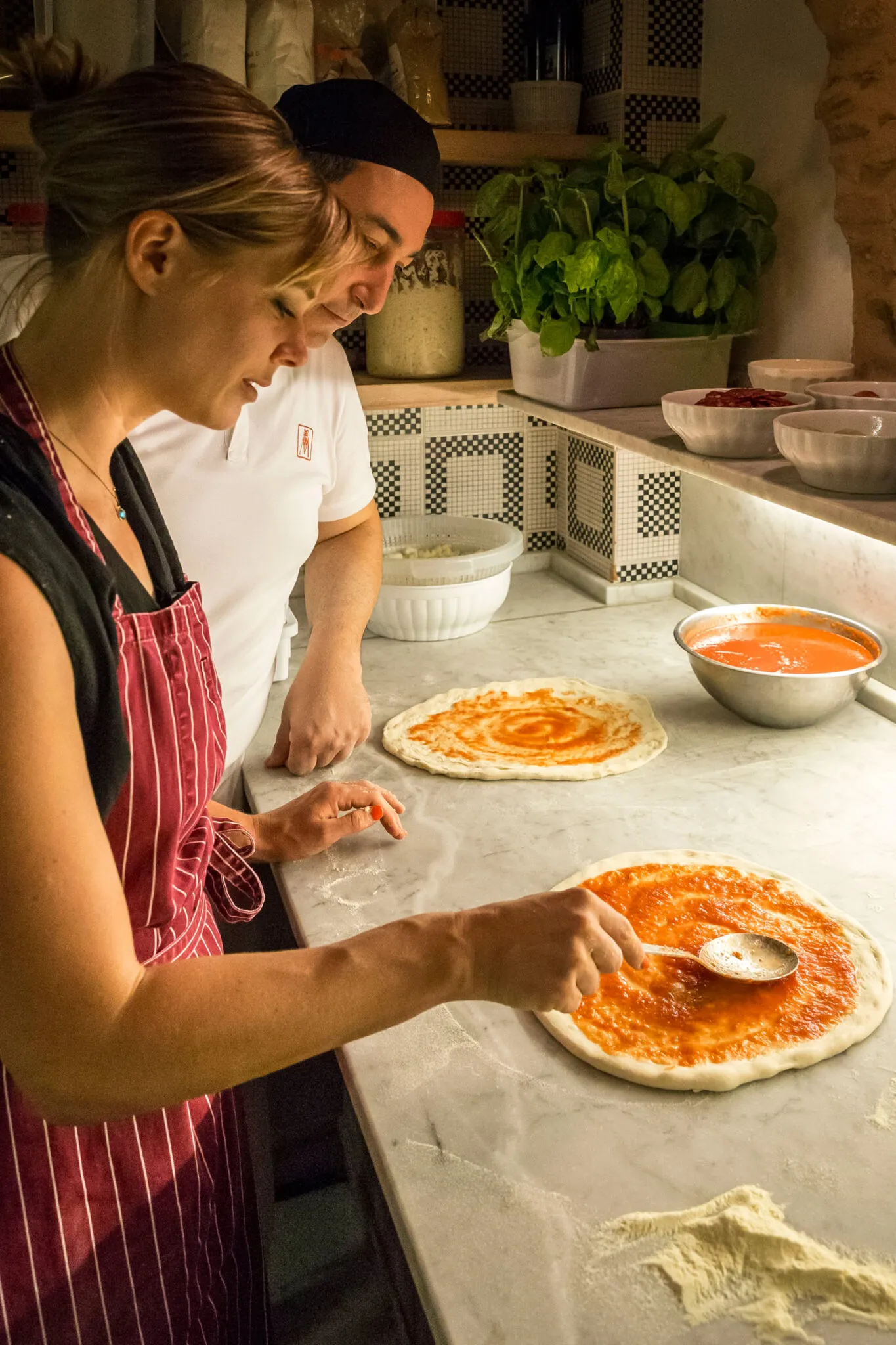 A woman in a red apron spreads sauce onto pizza dough