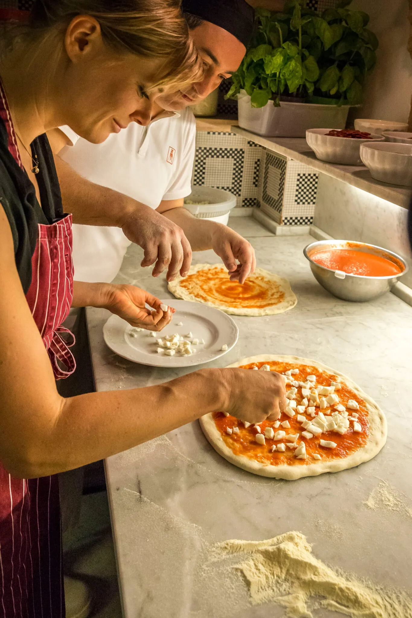 A woman tops a pizza with mozzarella while a chef watches