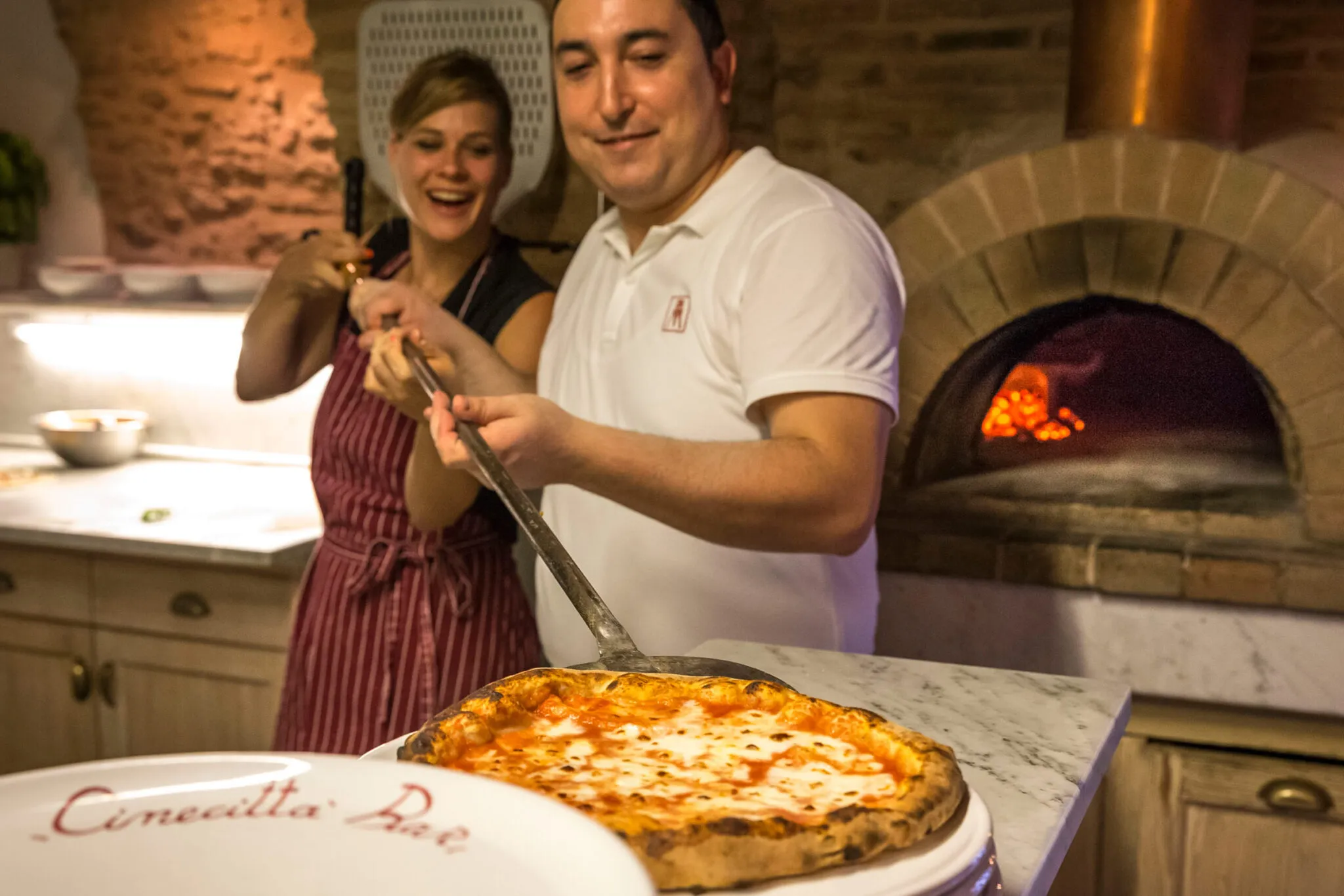 A chef helps a woman pull a hot pizza from a brick oven